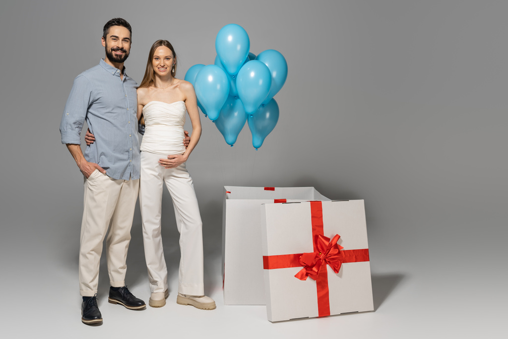 Parents-to-be posing beside a box revealing blue balloons at a gender reveal party.